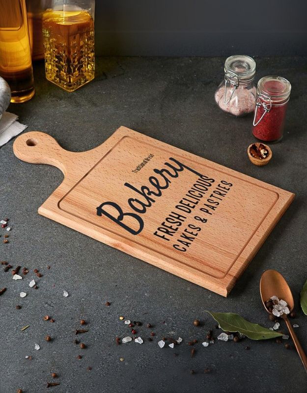 A rectangular acacia wood bread tray with a handle, serving as a cutting board. It has 'Bakery Fresh' printed on it, alongside decorative elements like spices and a glass of beer in the background.